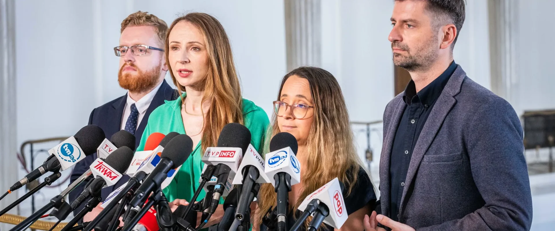 Kobieta przemawia do zestawu mikrofonów mediów podczas konferencji prasowej, w towarzystwie innych osób, w kontekście politycznym dotyczącym likwidacji małych porodówek w Polsce pod rządami PiS.