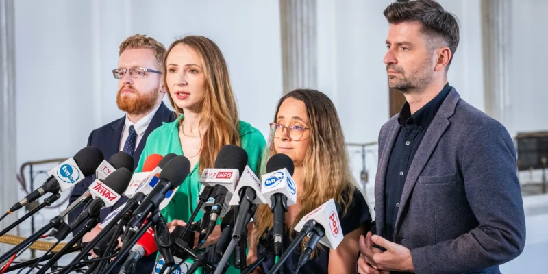 Kobieta przemawia do zestawu mikrofonów mediów podczas konferencji prasowej, w towarzystwie innych osób, w kontekście politycznym dotyczącym likwidacji małych porodówek w Polsce pod rządami PiS.