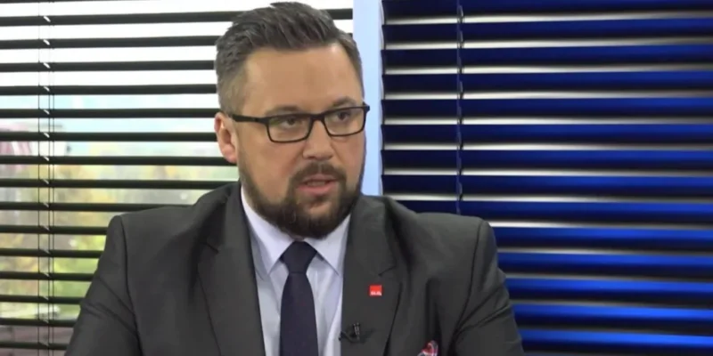 Mężczyzna w garniturze i okularach w studiu telewizyjnym, rozmawiający o konieczności zmiany rządu.