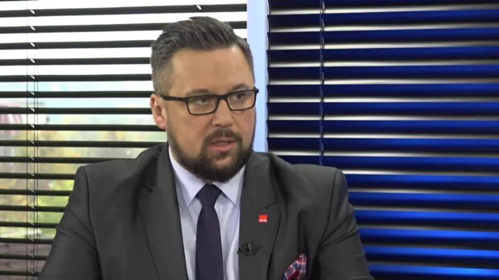 Mężczyzna w garniturze i okularach w studiu telewizyjnym, rozmawiający o konieczności zmiany rządu.