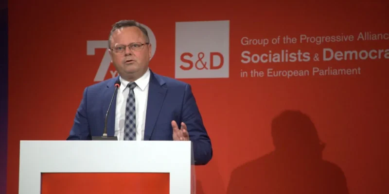 Mężczyzna w granatowym garniturze przemawia z mównicy na czerwonym tle z logo S&D oraz napisem o Grupie Socjalistów i Demokratów w Parlamencie Europejskim.