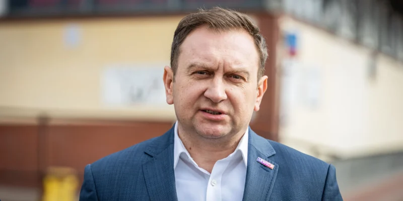 Mężczyzna w granatowym garniturze i białej koszuli stoi na zewnątrz, na klapie ma przypinkę Lewica, a w tle widać budynek.