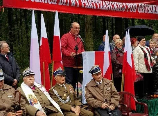 Weterani w mundurach siedzą na pierwszym planie, a na mównicy przemawia mężczyzna przed polskimi flagami i czerwonym banerem informującym o 73. rocznicy Bitwy pod Gruszką.