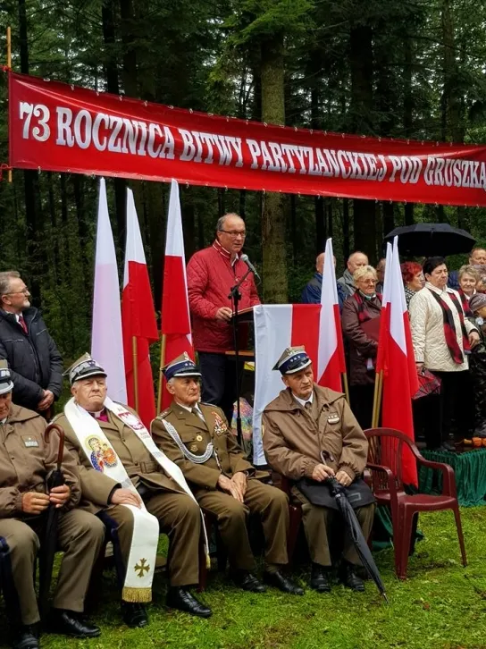Weterani w mundurach siedzą na pierwszym planie, a na mównicy przemawia mężczyzna przed polskimi flagami i czerwonym banerem informującym o 73. rocznicy Bitwy pod Gruszką.