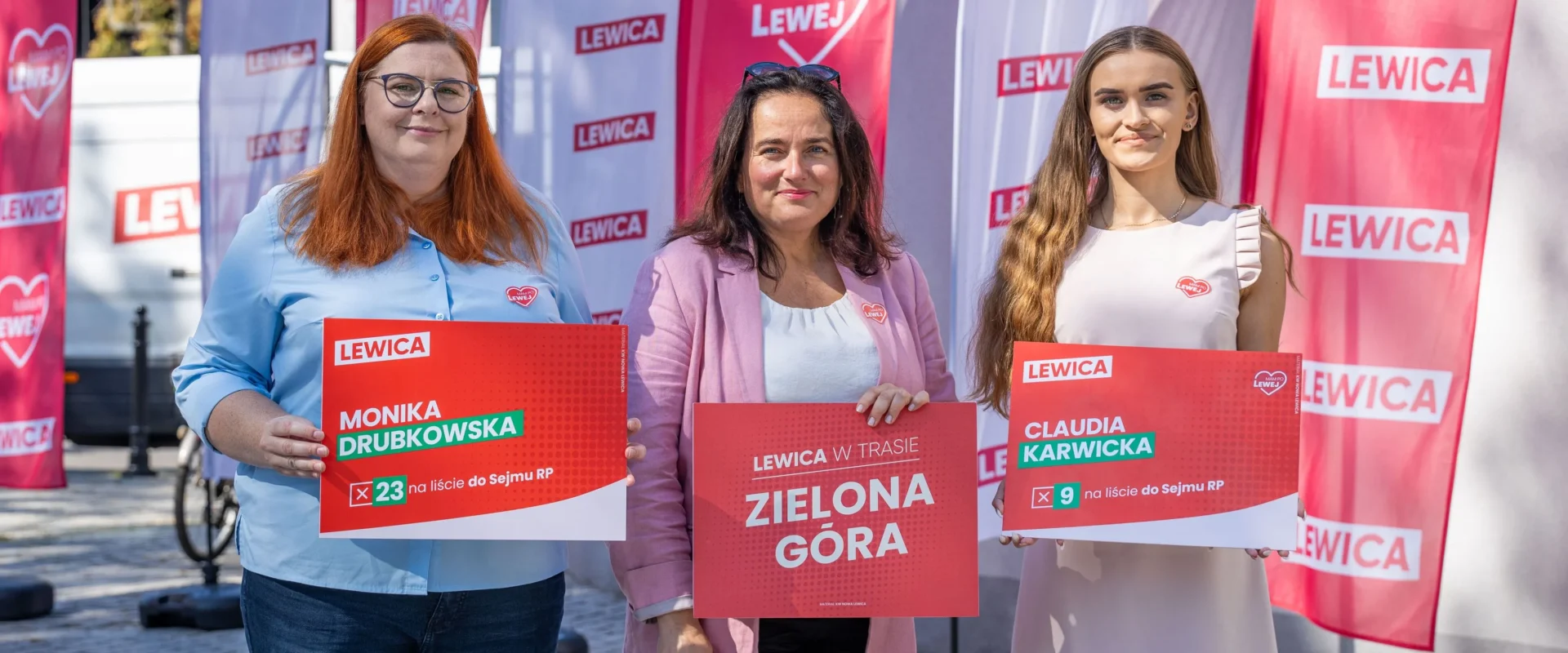 Trzy kobiety stoją na zewnątrz, każda trzyma plakat wyborczy Lewicy, a w tle widoczne czerwone flagi z napisem LEWICA.