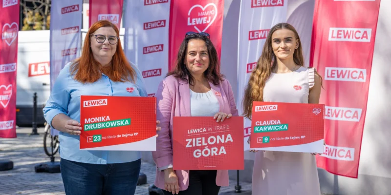 Trzy kobiety stoją na zewnątrz, każda trzyma plakat wyborczy Lewicy, a w tle widoczne czerwone flagi z napisem LEWICA.