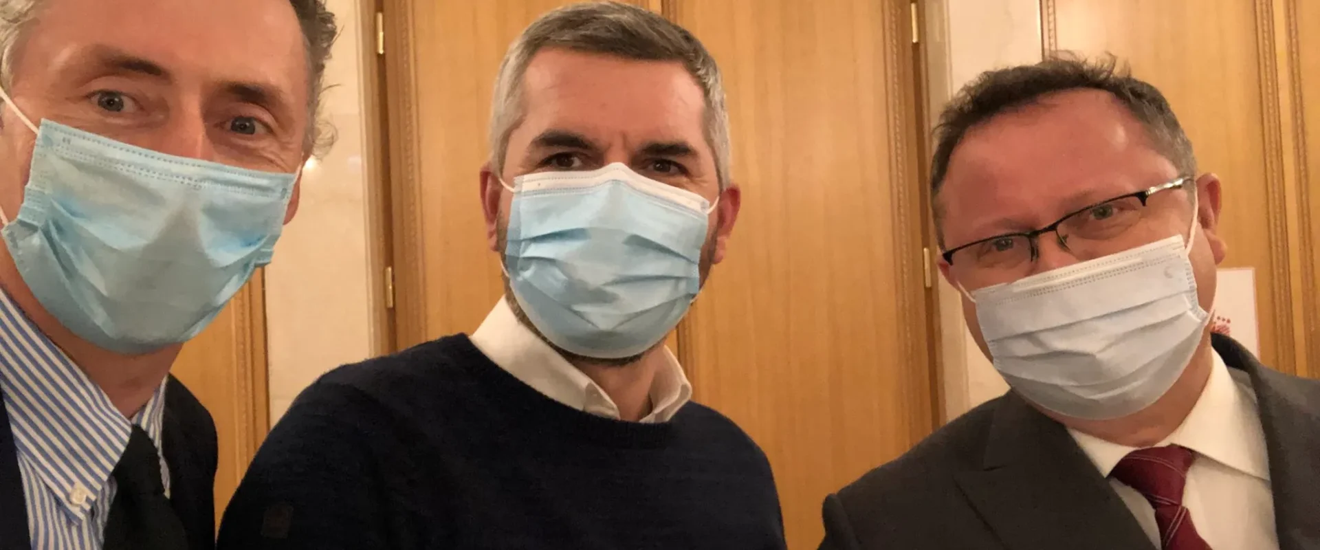 Trzech mężczyzn w maseczkach ochronnych stoi blisko siebie w wnętrzu budynku.