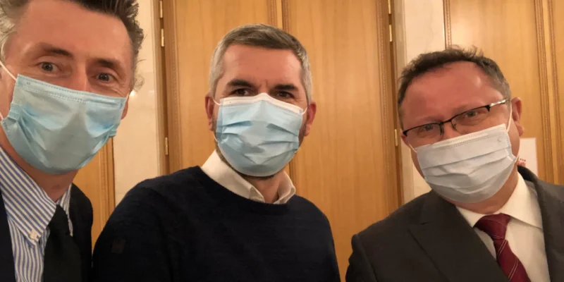 Trzech mężczyzn w maseczkach ochronnych stoi blisko siebie w wnętrzu budynku.