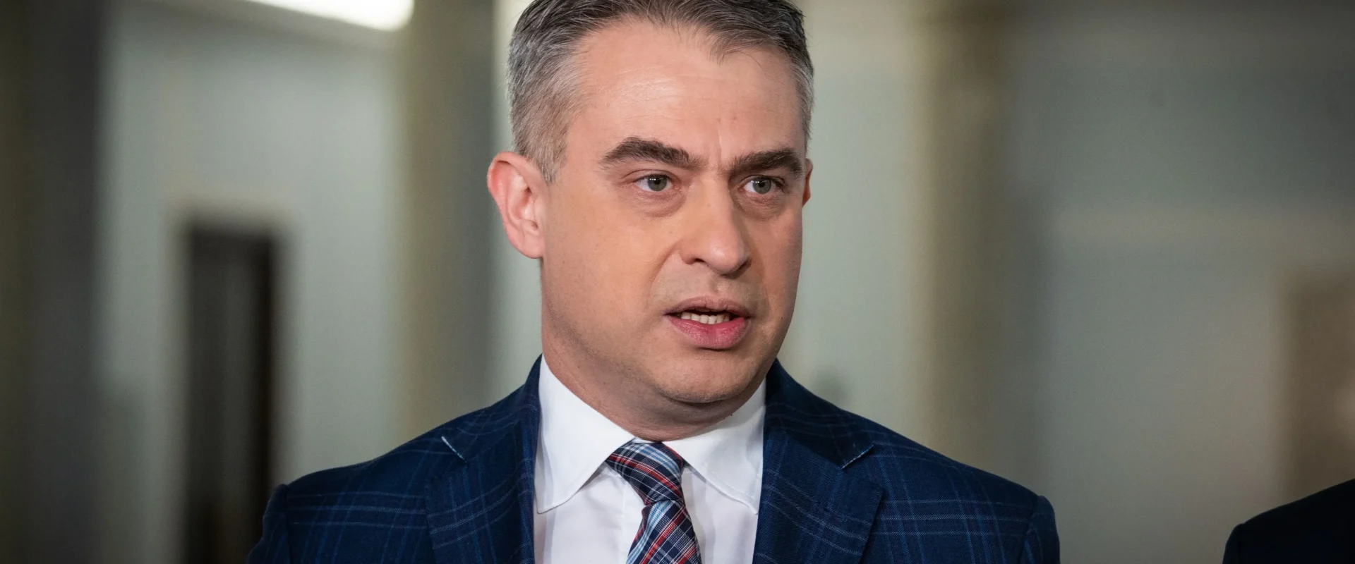 Polityk w granatowym garniturze z krawatem w kratę przemawia do mediów podczas konferencji prasowej.