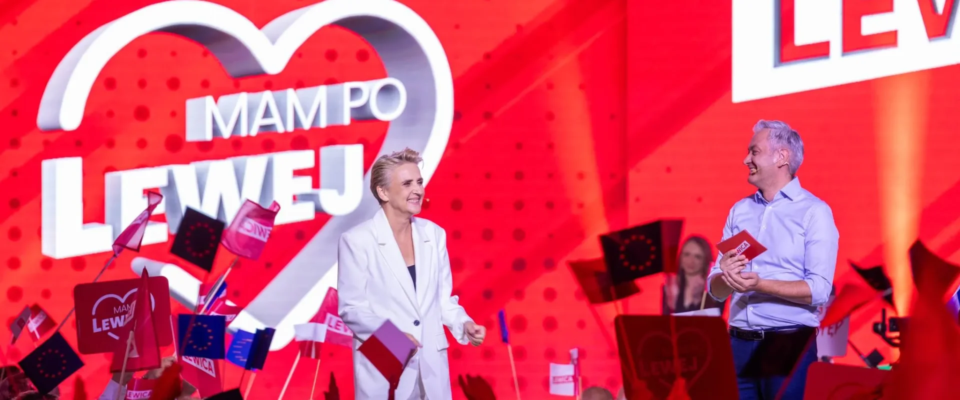 Na scenie podczas wiecu Lewicy dwie osoby prowadzą rozmowę — kobieta w białym garniturze i mężczyzna w błękitnej koszuli — wokół tłum z flagami, a w tle duży czerwony ekran z napisem LEWICA i logo w kształcie serca z napisem MAM PO LEWEJ.