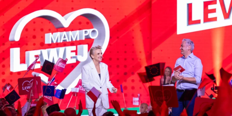 Na scenie podczas wiecu Lewicy dwie osoby prowadzą rozmowę — kobieta w białym garniturze i mężczyzna w błękitnej koszuli — wokół tłum z flagami, a w tle duży czerwony ekran z napisem LEWICA i logo w kształcie serca z napisem MAM PO LEWEJ.