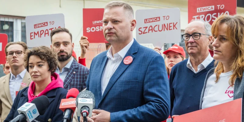 Grupa polityków Lewicy podczas konferencji prasowej, stojąca za mikrofonami, trzyma plakaty z hasłami „LEWICA TO EMPATIA”, „LEWICA TO SZACUNEK” i „LEWICA TO TROSKA”.