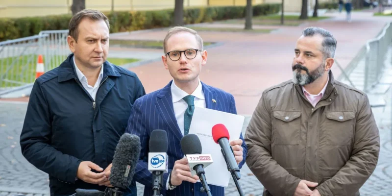 Na zdjęciu trzech mężczyzn w eleganckich ubraniach stoi przed mikrofonami, jeden z nich wygłasza przemówienie, a w tle widać ulicę z drzewami.