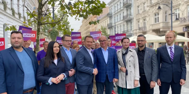 Grupa polityków Lewicy w marynarkach stoi na ulicy, w tle widoczne plakaty z napisem LEWICA, nawiązujące do zakończenia procesu konsolidacji przed kongresem zjednoczeniowym.