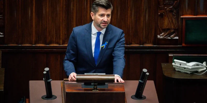 Mężczyzna w granatowym garniturze i niebieskim krawacie stoi za mównicą w sali plenarnej, z żółto-niebieską wstążką przypiętą do klatki piersiowej.