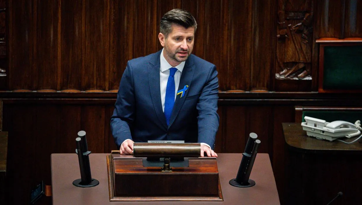 Mężczyzna w granatowym garniturze i niebieskim krawacie stoi za mównicą w sali plenarnej, z żółto-niebieską wstążką przypiętą do klatki piersiowej.