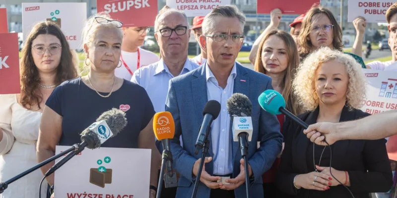 Grupa działaczy Lewicy na konferencji prasowej z plakatami i mikrofonami; w centrum przemawia mężczyzna w granatowym garniturze, otoczony innymi uczestnikami z hasłami o ochronie zdrowia i wyższych płac.