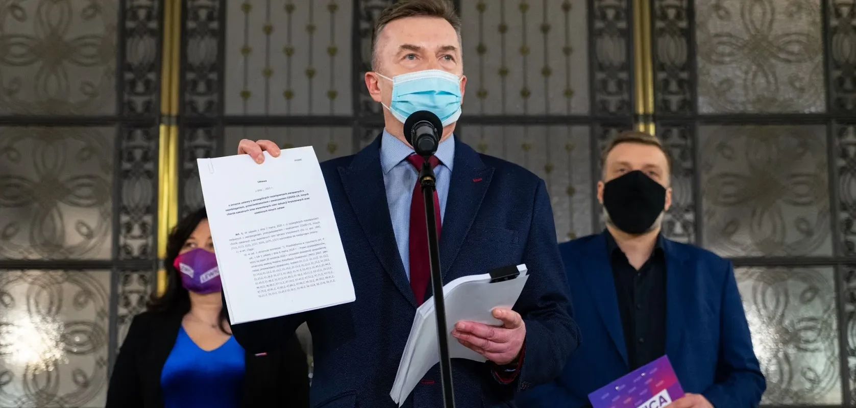 Polityk w garniturze i maseczce wygłasza oświadczenie z mikrofonem, trzymając dokumenty podczas konferencji Lewicy dotyczącej planu ratunkowego dla gospodarki.