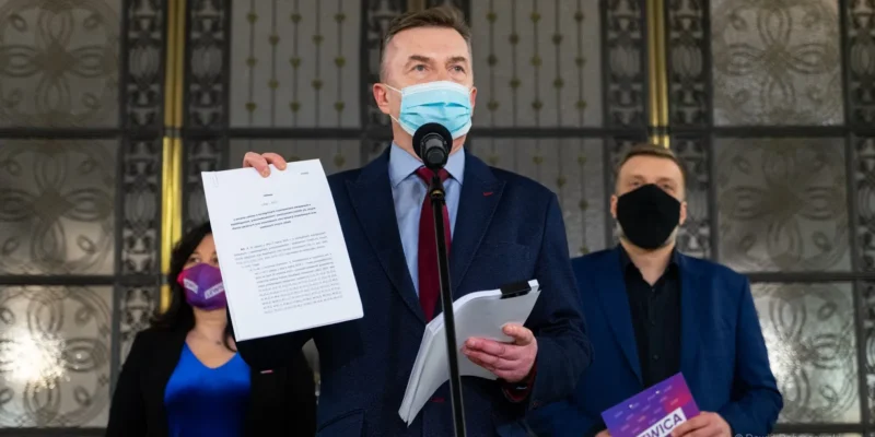 Polityk w garniturze i maseczce wygłasza oświadczenie z mikrofonem, trzymając dokumenty podczas konferencji Lewicy dotyczącej planu ratunkowego dla gospodarki.