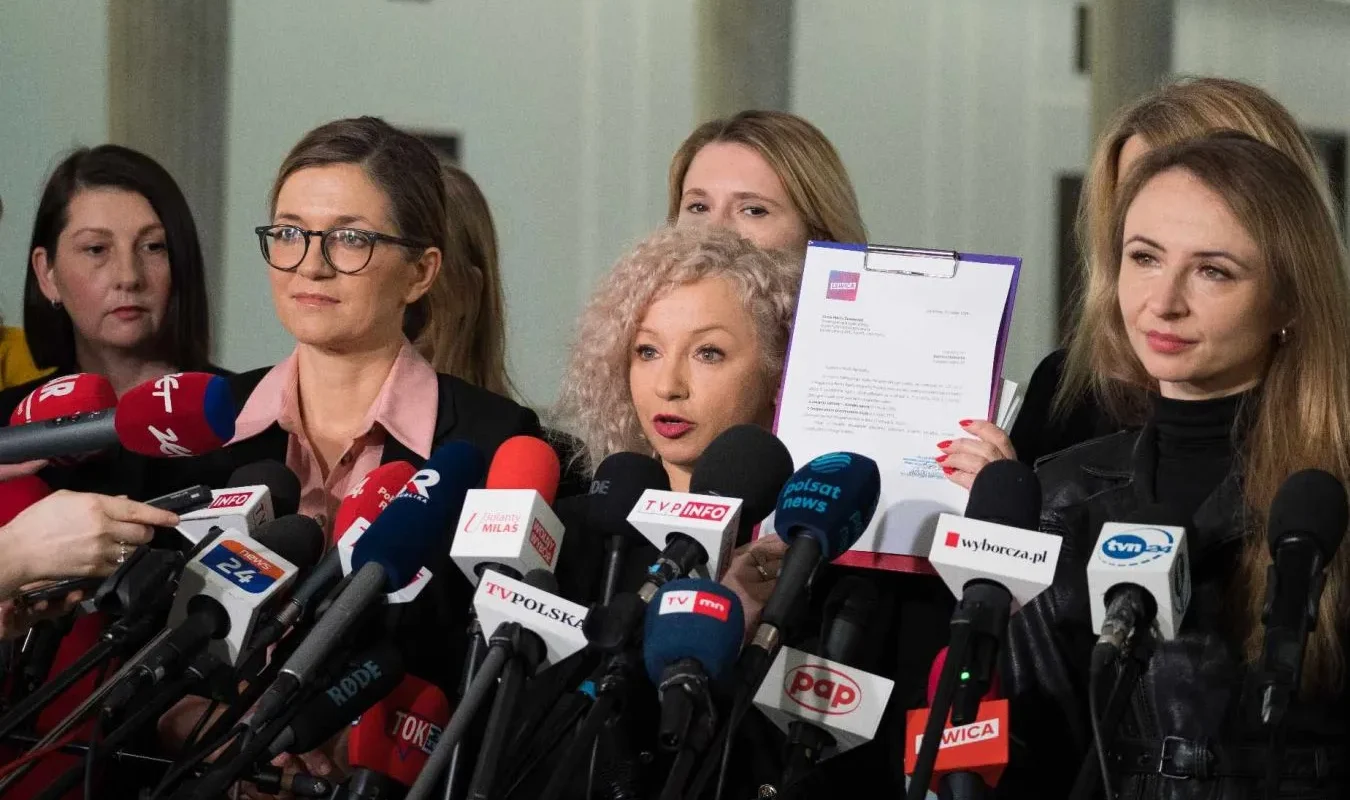 Grupa kobiet podczas konferencji prasowej otoczona licznymi mikrofonami mediów, jedna z nich trzyma dokumenty.