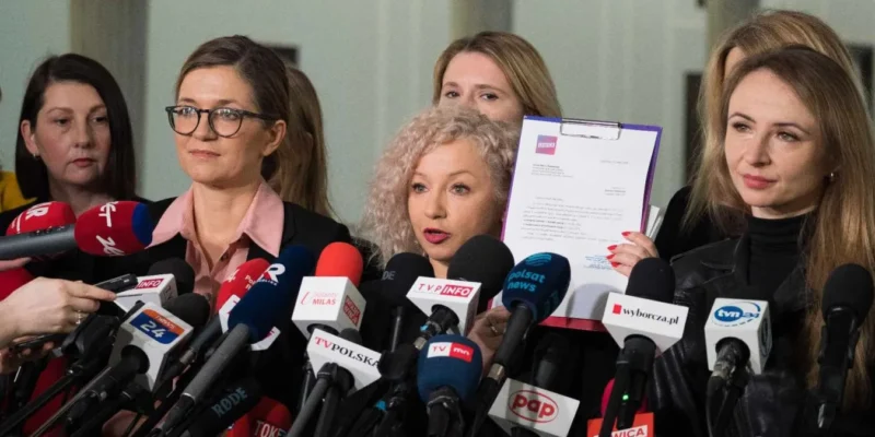Grupa kobiet podczas konferencji prasowej otoczona licznymi mikrofonami mediów, jedna z nich trzyma dokumenty.