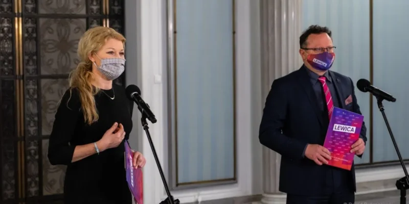 Dwóch polityków Lewicy w maseczkach stoi przy mikrofonach podczas konferencji prasowej, trzymając materiały z napisem Lewica, w tle kolumny i drzwi.