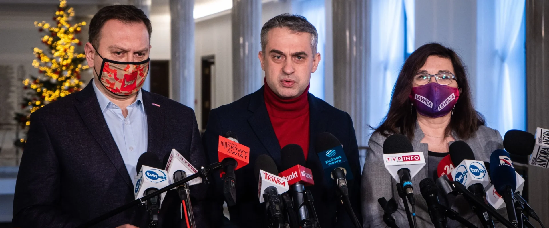 Trzech osób stoi za licznymi mikrofonami podczas konferencji prasowej; centralnie przemawia mężczyzna w czerwonym golfie, po lewej mężczyzna w maseczce z czerwono-pomarańczowym wzorem, a po prawej kobieta w maseczce z napisem Lewica, w tle kolumny i dekorowana choinka.