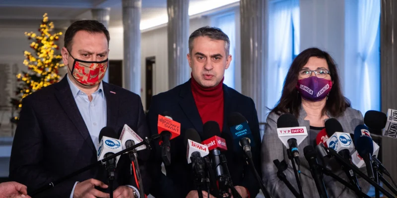 Trzech osób stoi za licznymi mikrofonami podczas konferencji prasowej; centralnie przemawia mężczyzna w czerwonym golfie, po lewej mężczyzna w maseczce z czerwono-pomarańczowym wzorem, a po prawej kobieta w maseczce z napisem Lewica, w tle kolumny i dekorowana choinka.