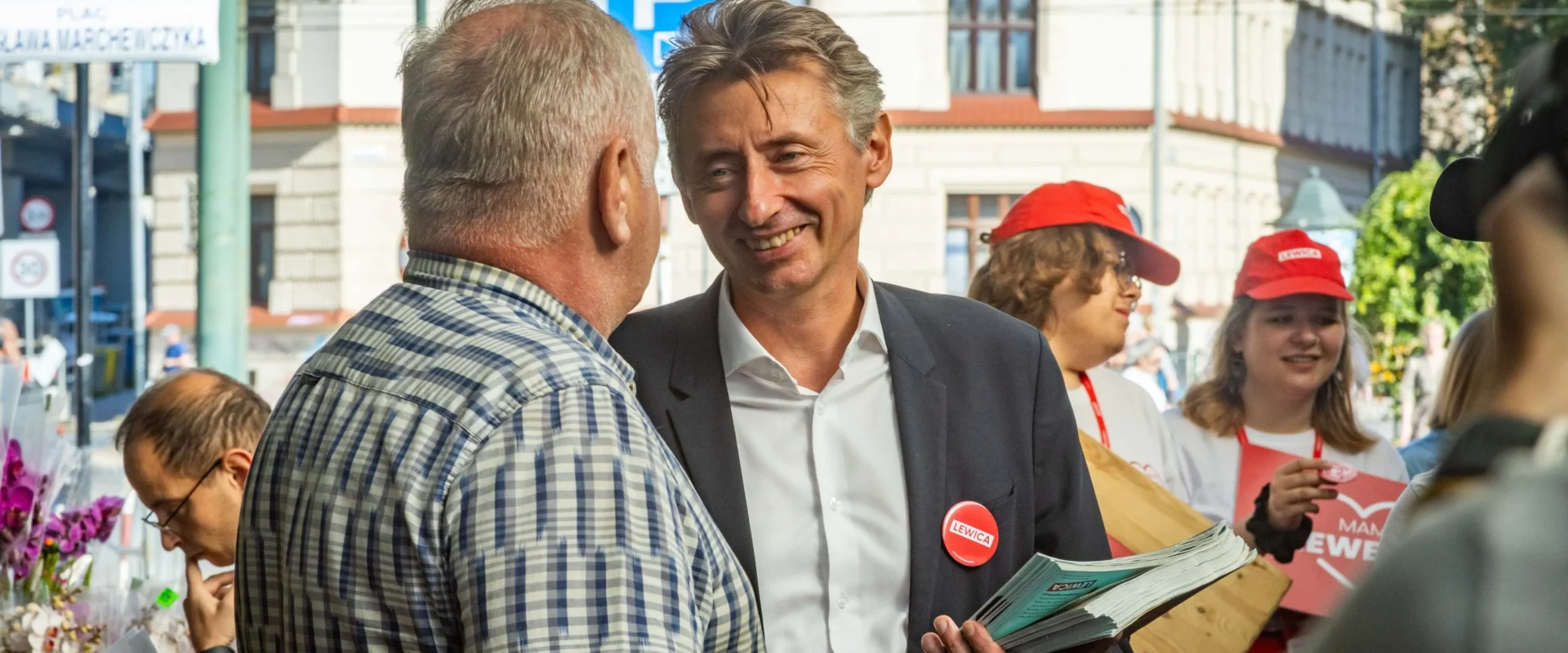 Uśmiechnięty mężczyzna w garniturze rozdaje gazety i rozmawia z innym mężczyzną na ulicy, w tle widać wolontariuszy w czerwonych czapkach z napisem Lewica.