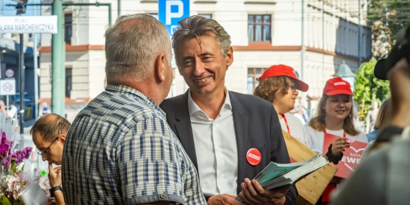 Uśmiechnięty mężczyzna w garniturze rozdaje gazety i rozmawia z innym mężczyzną na ulicy, w tle widać wolontariuszy w czerwonych czapkach z napisem Lewica.