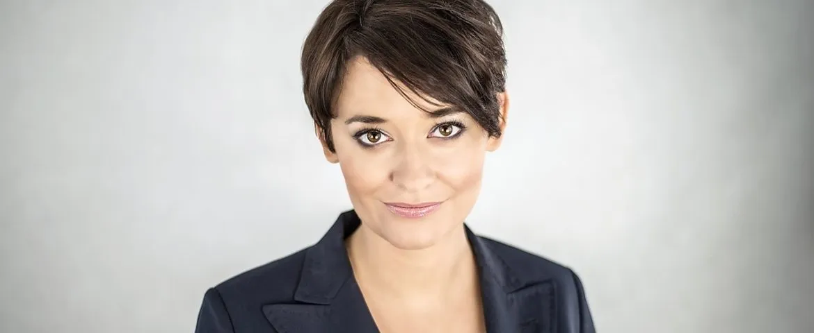 Portret uśmiechniętej kobiety o krótkich ciemnych włosach w granatowej marynarce na jasnym, rozmytym tle.