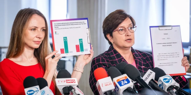 Na konferencji prasowej dwie kobiety prezentują materiały o klauzuli sumienia: jedna trzyma planszę z wykresami, druga kartę z projektem ustawy, przed licznymi mikrofonami.