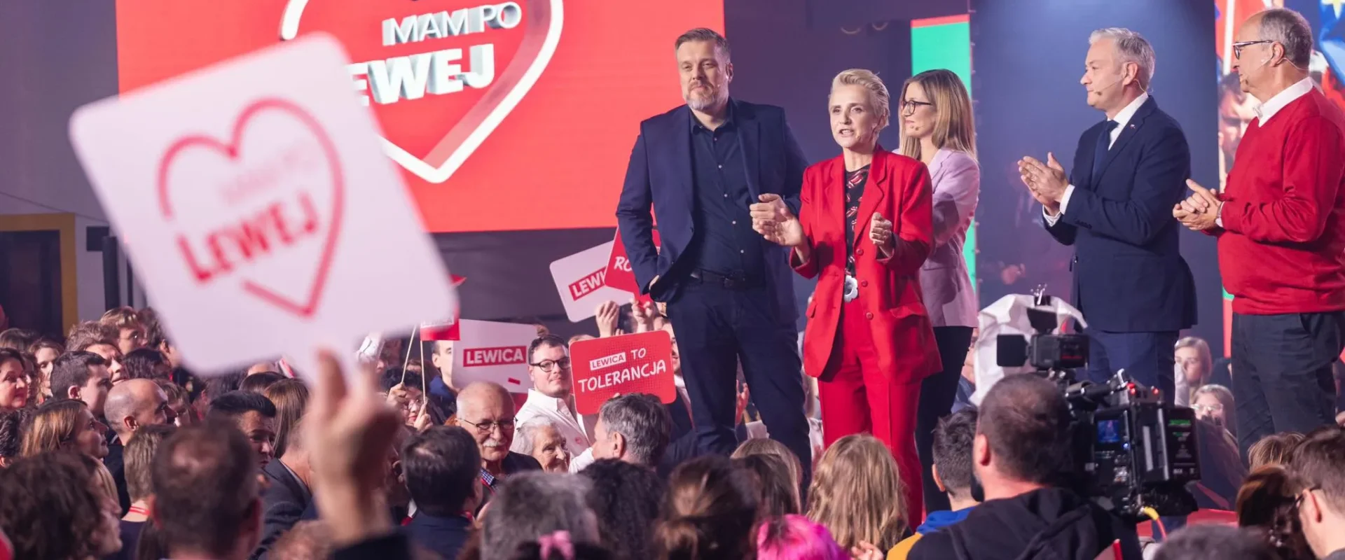 Podczas wieczoru wyborczego grupa polityków stoi na scenie wśród wiwatującego tłumu z plakatami Lewica, na tle dużego czerwonego ekranu z sercem.