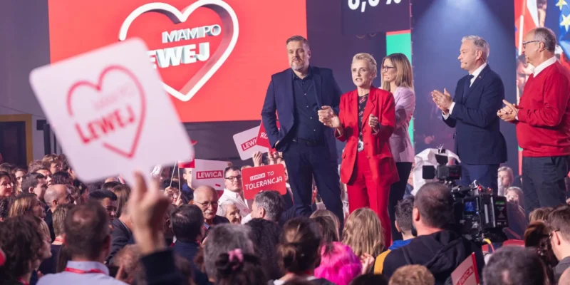 Podczas wieczoru wyborczego grupa polityków stoi na scenie wśród wiwatującego tłumu z plakatami Lewica, na tle dużego czerwonego ekranu z sercem.