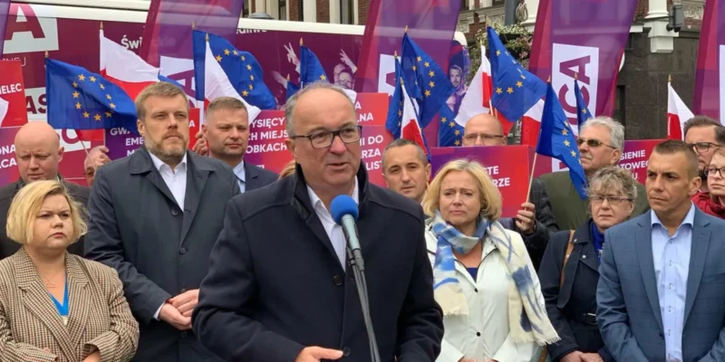 Mężczyzna z okularami przemawia do mikrofonu podczas wiecu lewicowego, w tle tłum ludzi, flagi UE i Polski oraz kolorowe banery.