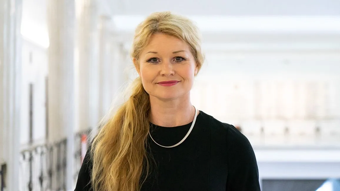 Uśmiechnięta kobieta o blond włosach związanych w koński ogon stoi w jasnym korytarzu.