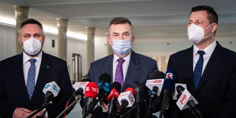 Trzej mężczyźni w maskach ochronnych stoją przed mikrofonami podczas konferencji prasowej w jasnym wnętrzu.