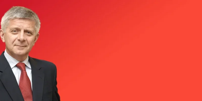 Portret mężczyzny w czarnym garniturze, białej koszuli i czerwonym krawacie na jednolitym czerwonym tle.