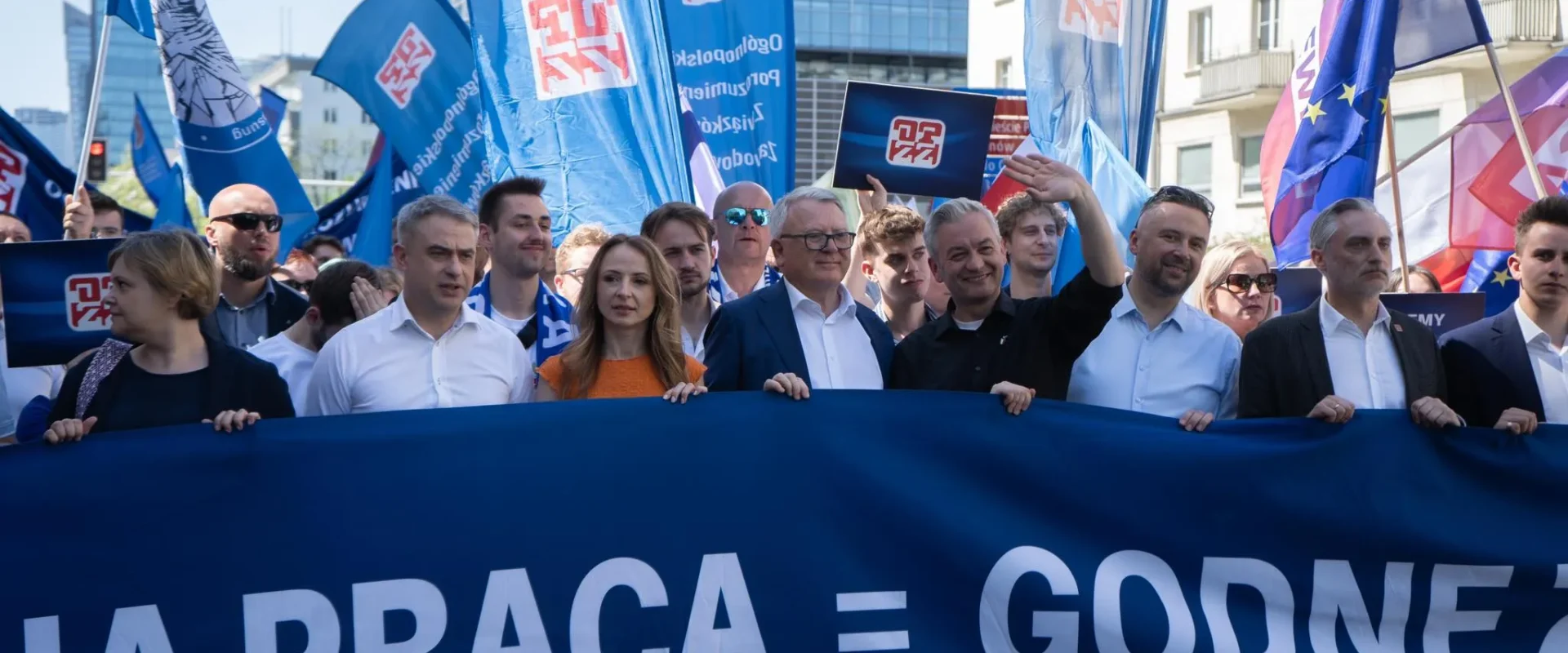 Grupa uczestników demonstracji prowadzi marsz wzdłuż ulicy podczas Święta Pracy 2024, trzymając duży niebieski baner z białym napisem i liczne flagi.