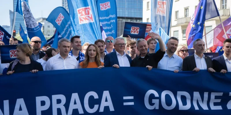Grupa uczestników demonstracji prowadzi marsz wzdłuż ulicy podczas Święta Pracy 2024, trzymając duży niebieski baner z białym napisem i liczne flagi.