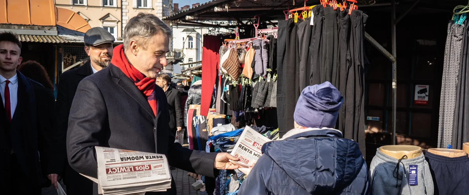 Na zdjęciu mężczyzna w eleganckim ubraniu, trzymający gazety, rozmawia z kobietą w ciepłej odzieży na stoisku na targu.