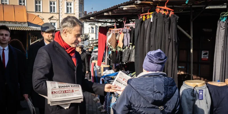 Na zdjęciu mężczyzna w eleganckim ubraniu, trzymający gazety, rozmawia z kobietą w ciepłej odzieży na stoisku na targu.