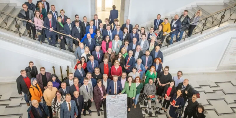 Duża grupa osób w formalnych ubraniach stoi na marmurowej klatce schodowej, tworząc zdjęcie grupowe pod hasłem „Silny samorząd to silna Polska”.