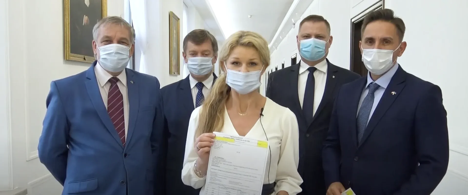 Grupa osób w garniturach i maseczkach stoi w białym korytarzu z obrazami na ścianach; przed nimi kobieta w białej bluzce trzyma dokumenty, a w rogu kadru widnieje logo Lewicy.