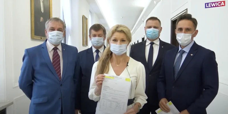 Grupa osób w garniturach i maseczkach stoi w białym korytarzu z obrazami na ścianach; przed nimi kobieta w białej bluzce trzyma dokumenty, a w rogu kadru widnieje logo Lewicy.