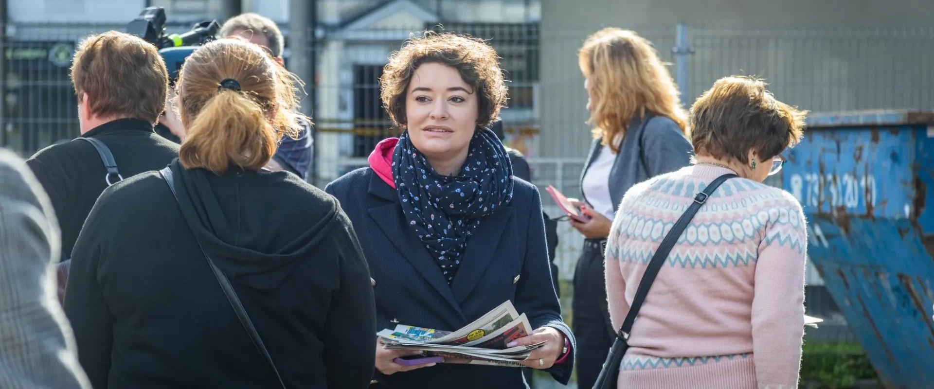 Kobieta w granatowym garniturze i niebieskim szalu trzyma gazety, stojąc wśród grupy ludzi na zewnątrz, z widocznym ogrodzeniem i kontenerem na odpady w tle.