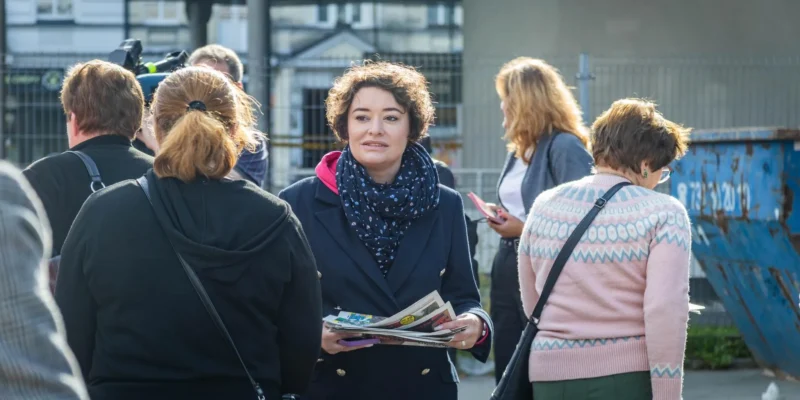 Kobieta w granatowym garniturze i niebieskim szalu trzyma gazety, stojąc wśród grupy ludzi na zewnątrz, z widocznym ogrodzeniem i kontenerem na odpady w tle.