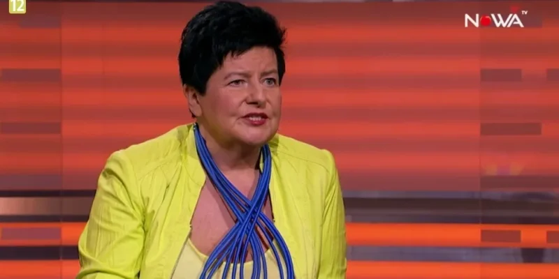 Kobieta w żółtej marynarce i efektownym niebieskim naszyjniku mówi w studiu telewizyjnym na tle czerwono‑pomarańczowych pasów.