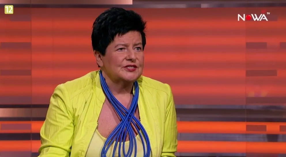 Kobieta w żółtej marynarce i efektownym niebieskim naszyjniku mówi w studiu telewizyjnym na tle czerwono‑pomarańczowych pasów.