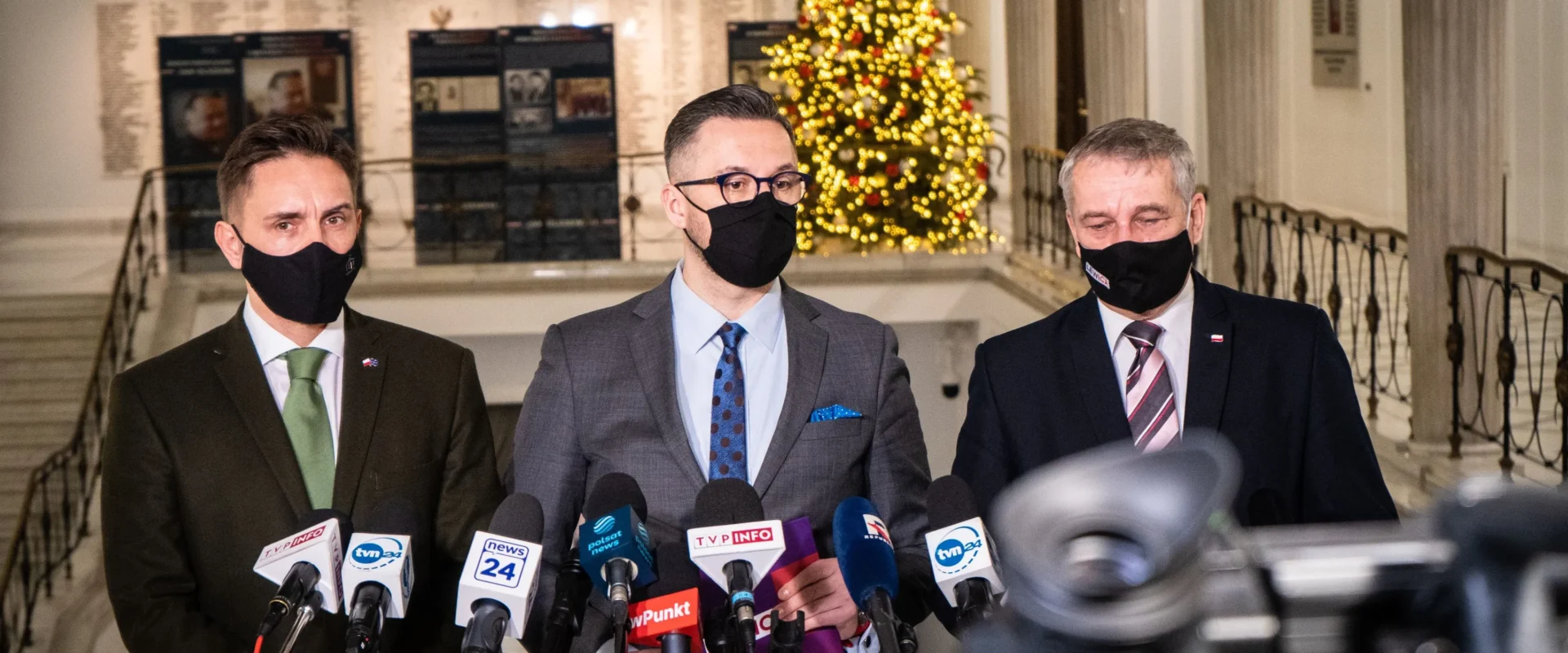 Trzej mężczyźni w eleganckich strojach, noszący maseczki, stoją przed mikrofonami w dekorowanej bożonarodzeniowo przestrzeni.